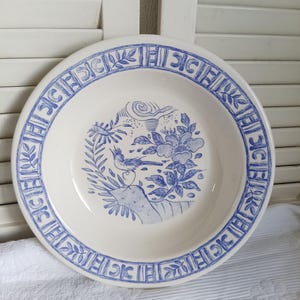Op de afbeelding: Een ronde keramische kom met een crèmekleurige basis en blauwe transferware-patronen. De rand is voorzien van een herhalende geometrische en bladvormige rand, terwijl het midden een gedetailleerde illustratie toont van een vogel op een rots.