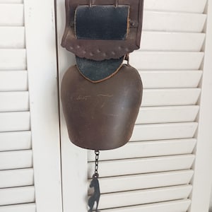 Peut inclure: Une cloche en métal brun foncé fixée à une sangle en cuir noir avec un support en cuir brun festonné. Une fine chaîne métallique pend au bas de la cloche, ornée d'une silhouette en métal noir représentant un chat en position assise.