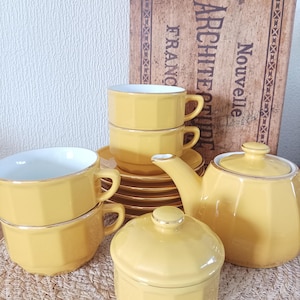 May include: A yellow tea set with gold edging, comprising cups, saucers, a teapot, and a sugar bowl. The set is displayed on a woven mat, with a vintage book in the background. The cups are stacked, and the teapot has a lid.