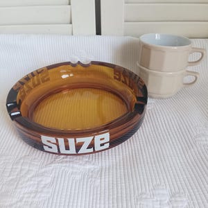 Peut inclure: Un cendrier rond en verre ambré avec le texte blanc SUZE imprimé sur le côté, doté de trois encoches sur le bord. À côté du cendrier se trouvent deux tasses en céramique beige empilées avec un design à facettes et des anses latérales.