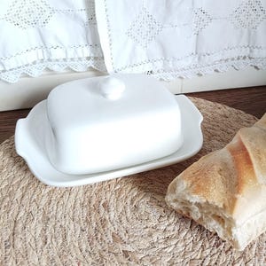 Peut inclure: Un beurrier en céramique blanche composé d'une base rectangulaire, d'un couvercle assorti et d'un petit bouton de préhension sur le dessus. L'objet présente des poignées latérales intégrées et une finition lisse et brillante.