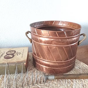 May include: A copper-coloured metal bucket with two handles, a vintage whisk, and a small, aged book. The bucket has a diagonal pattern and sits on a woven mat. The whisk has a wooden handle and a metal coil.