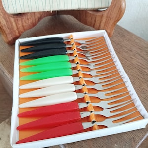 Può includere: Un set di dodici forchettine da antipasto in acciaio inossidabile disposte in una scatola bianca con inserto arancione. Le forchette hanno manici affusolati neri, verdi, bianchi e rossi. Il coperchio riporta la scritta Little Forks STAINLESS STEEL.