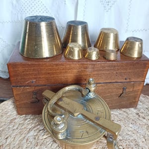 Peut inclure: Ensemble de poids anciens en laiton de différentes tailles et une balance en laiton. Les poids sont en forme de cône et sont posés sur une boîte en bois. La balance est ronde avec une poignée et un levier.