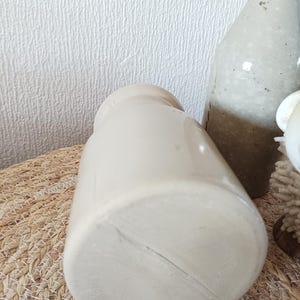 May include: A cylindrical, off-white ceramic vessel with a smooth, glazed finish and a narrow neck. The base is flat and circular, showing a subtle linear mark across the surface. The vessel is positioned upright, displaying a simple, uniform design.