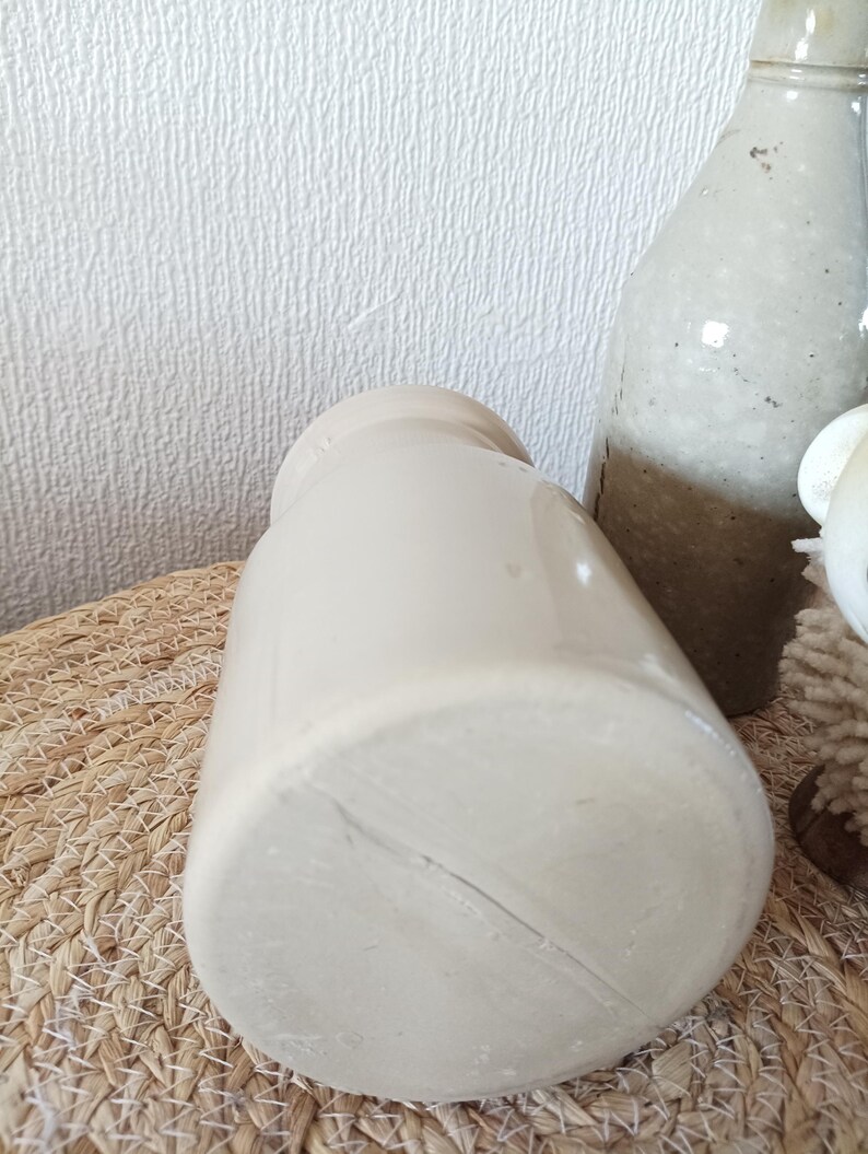 May include: A cylindrical, off-white ceramic vessel with a smooth, glazed finish and a narrow neck. The base is flat and circular, showing a subtle linear mark across the surface. The vessel is positioned upright, displaying a simple, uniform design.