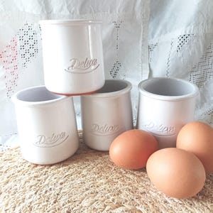 Peut inclure: Quatre pots en céramique blanche avec une finition lisse, chacun portant le mot Deluxe en relief sur le devant. Les pots sont cylindriques avec un bord légèrement évasé. Trois œufs bruns sont placés devant les pots sur une surface tressée.