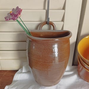 Peut inclure: Un vase mural en céramique marron avec une poignée et un crochet en métal pour l'accrocher. Une fleur violette et rose avec des tiges vertes est à l'intérieur du vase. Le vase est posé sur un tissu blanc.