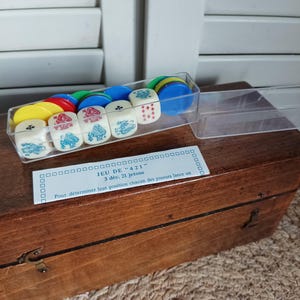 Op de afbeelding: Een vintage houten speldoos met een doorzichtige plastic container met dobbelstenen en kleurrijke fiches. De dobbelstenen zijn wit met blauwe illustraties. Op de doos staat "JEU DE '421'".