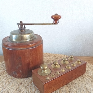 Pode incluir: Um antigo moedor de café de madeira com tampa e manivela de metal, ao lado de uma caixa de madeira contendo pesos de latão. O moedor tem uma base cilíndrica e uma manivela de madeira. Os pesos são de vários tamanhos.