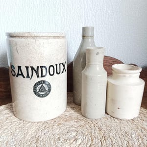 May include: Four antique stoneware containers in varying shapes and sizes. The largest is a cylindrical jar with the word "SAINDOUX" in black lettering. The other three are bottles and jars in shades of beige and off-white.