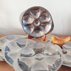 May include: A set of vintage stainless steel oyster serving platters. Each platter has six rounded compartments and a central well. A small, ceramic bird figurine is also present.