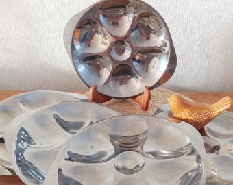 Puede incluir: Un juego de fuentes vintage de acero inoxidable para servir ostras. Cada fuente tiene seis compartimentos redondeados y un pozo central. También hay una pequeña figura de pájaro de cerámica.