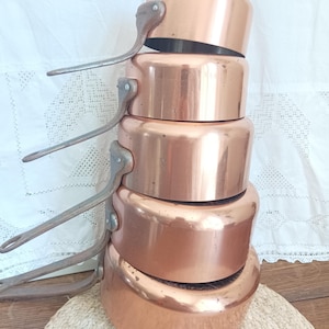 Peut inclure: Une pile de cinq casseroles en cuivre avec des poignées en métal rivetées. Les casseroles sont d'un cuivre brillant et chaud, empilées les unes sur les autres. Les poignées sont de couleur argent.
