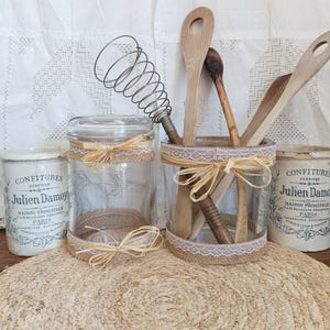 Peut inclure: Deux bocaux en verre transparent décorés de toile de jute et de dentelle blanche, chacun noué avec un ruban de raphia. Un bocal contient un fouet en métal et trois cuillères en bois. Deux pots en céramique blanche Confitures Julien Damoy.