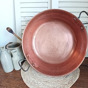 Peut inclure: Une grande poêle en cuivre antique avec deux poignées noires. La poêle est ronde avec un bol large et peu profond. Elle repose sur un dessous de plat circulaire tissé. Une cuillère en bois et plusieurs bouteilles en céramique sont en arrière-plan.