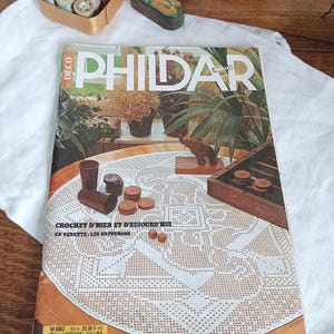 Può includere: Una rivista vintage intitolata "PHILDAR" con un motivo all'uncinetto sulla copertina. La copertina presenta una tovaglia all'uncinetto bianca, pedine di legno e una piccola statuina di elefante in legno. Altri oggetti includono rocchetti di filo e una piccola latta.