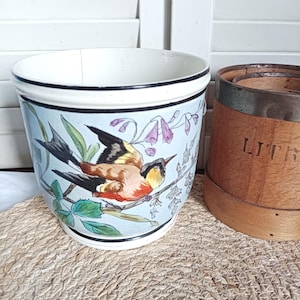 Może przedstawiać: Ceramiczna donica z motywem ptaków i kwiatów. Donica ma jasnoniebieskie tło, czarną obwódkę i białe wnętrze. Obok donicy znajduje się drewniany pojemnik.