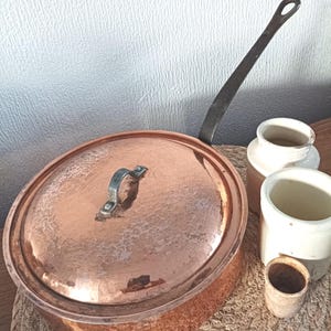 Op de afbeelding: Een ronde koperen pan met een gehamerde textuur, een bijpassend deksel met een metalen handvat en een lange, donkere metalen steel aan de zijkant. De pan wordt vergezeld door drie kleine, cilindrische, gebroken witte keramische bakjes van verschillende grootte.