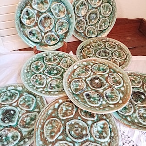 Peut inclure: Ensemble d'assiettes à huîtres en céramique, dans des tons de turquoise, marron et crème. Chaque assiette présente des creux moulés pour les huîtres. Les assiettes sont disposées sur une nappe blanche et une surface en bois.