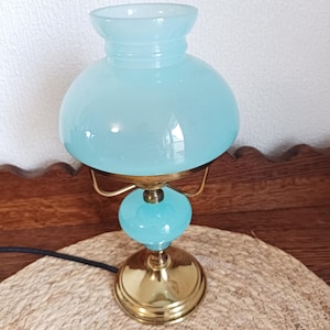 Könnte beinhalten: Eine Tischlampe im Vintage-Stil mit hellblauem Glasschirm und -kugel. Die Lampe hat einen Messingfuß und -akzente sowie ein schwarzes Netzkabel. Die Lampe steht auf einer gewebten Matte.