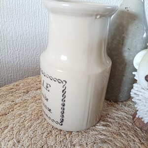 May include: A cream-coloured ceramic pitcher with a flared rim and a cylindrical body. The front features black printed text enclosed in a decorative rope-style border. The pitcher has a smooth, glossy finish and a slightly tapered neck design.