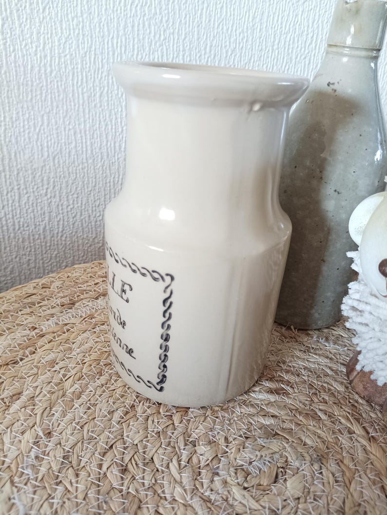 May include: A cream-coloured ceramic pitcher with a flared rim and a cylindrical body. The front features black printed text enclosed in a decorative rope-style border. The pitcher has a smooth, glossy finish and a slightly tapered neck design.