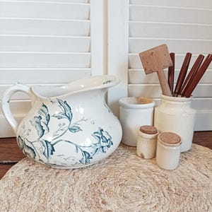 Peut inclure: Nature morte de style vintage avec une cruche blanche ornée de motifs floraux bleus, de petits pots en céramique avec des couvercles en liège et des ustensiles en bois. L'ensemble est disposé sur un tapis tressé.