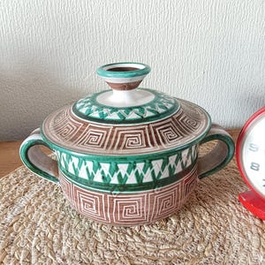 Op de afbeelding: Een keramische serveerschaal met deksel met een bruin, groen en wit geometrisch patroon. De schaal heeft twee handvatten en een kleine knop op het deksel. De schaal staat op een geweven placemat.