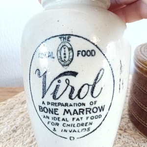 May include: Cream-coloured ceramic jar with a rounded body and a slightly flared rim. The jar has a black circular label with the word "Virol" in script and text that reads "A Preparation of Bone Marrow".