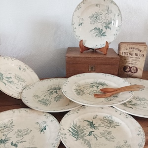 May include: A set of vintage white plates featuring a green floral pattern. The plates are displayed on a wooden surface, with a wooden box and a book in the background. Two wooden forks rest on one plate.