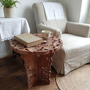 May include: A detailed, carved wooden side table with floral designs. A vintage book and a floral teacup sit on the table. The table is brown and stands on a wooden floor. A fern in a pot and a linen armchair are in the background.