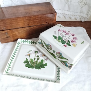 Peut inclure: Une boîte en bois et deux plats en céramique blanche avec des motifs floraux. Un plat carré avec une bordure de feuilles vertes et une illustration de marguerite. L'autre est un beurrier rectangulaire avec un motif floral et le texte "Cyclamen Repandum".