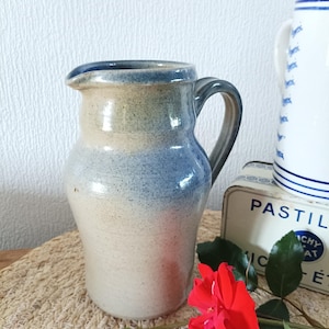May include: A handcrafted ceramic jug with a blue and beige colour gradient. The jug features a curved handle and a pouring spout. A red rose and a decorative tin are in the foreground.