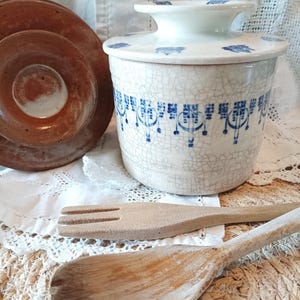 Puede incluir: Una mantequera de cerámica con esmalte blanco craquelado y patrones geométricos azules, con tapa a juego. Junto a ella hay un cuenco de cerámica marrón, un tenedor de madera y una cuchara de madera. La mantequera tiene un cuerpo cilíndrico.