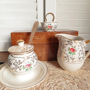Peut inclure: Un service à thé vintage de couleur crème avec des bordures dorées et des motifs floraux. L'ensemble comprend un sucrier avec couvercle, une crémière et un petit bol avec une anse. Une cuillère en bois est dans le sucrier. L'ensemble est présenté sur un tapis tressé.