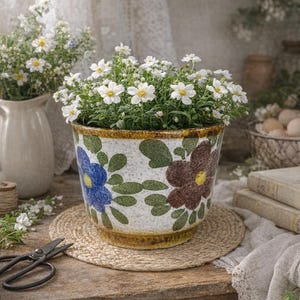 Op de afbeelding: Een keramische bloempot met een bloemenontwerp, gevuld met witte bloemen met gele centra. De pot heeft blauwe en bruine bloemen met groene bladeren. Hij staat op een ronde geweven mat op een houten tafel. Een vaas met witte bloemen staat op de achtergrond.