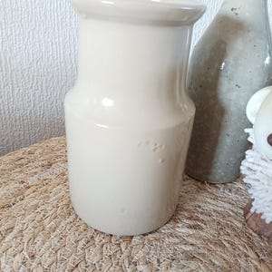 May include: A smooth, cream-coloured ceramic pitcher with a wide, flared rim and a cylindrical body. The surface features a glossy finish with subtle indentations on the side. The vessel stands upright with a flat base and a simple, clean design.
