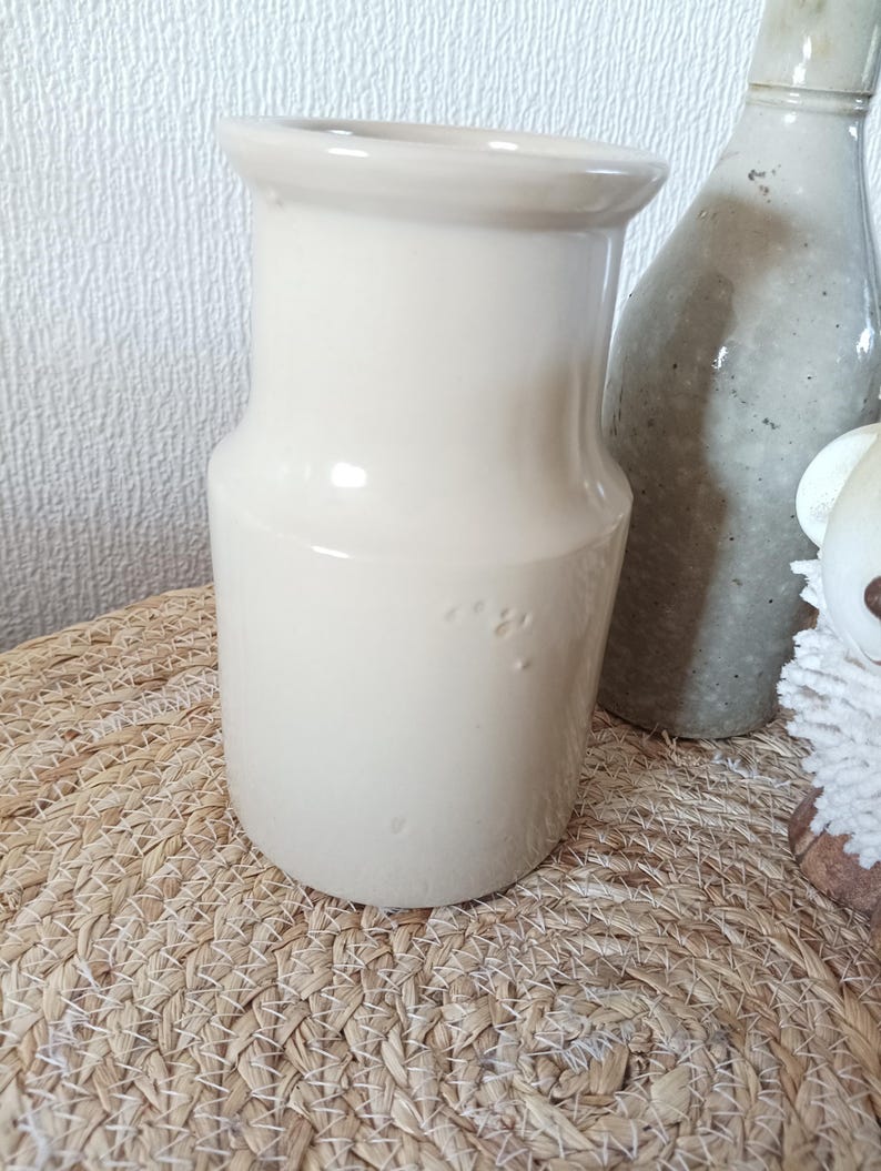 May include: A smooth, cream-coloured ceramic pitcher with a wide, flared rim and a cylindrical body. The surface features a glossy finish with subtle indentations on the side. The vessel stands upright with a flat base and a simple, clean design.