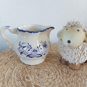May include: A white ceramic creamer with blue floral patterns and a scalloped rim. The creamer has a curved handle and a spout. A decorative sheep figurine is next to the creamer. The creamer is on a woven mat.