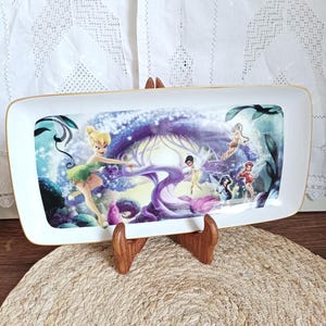 Puede incluir: Bandeja rectangular con borde dorado, con una colorida ilustración de hadas en un entorno forestal caprichoso. La bandeja está sostenida por un soporte de madera y descansa sobre un mantel individual tejido. El fondo es un mantel de encaje blanco.