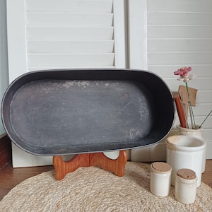Op de afbeelding: Een donkergrijze, ovale metalen schaal, mogelijk voor het serveren, staat op een houten standaard. De schaal heeft een licht versleten uiterlijk. Twee kleine, witte keramische potjes met kurken deksels staan vooraan.