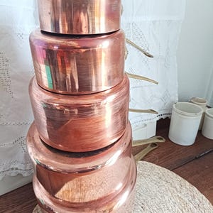 May include: A stack of four copper pots with lids, showcasing a warm, reddish-brown hue. The pots are arranged in a nested fashion, with brass handles visible on the sides. The cookware sits on a woven placemat, suggesting a kitchen or dining setting.