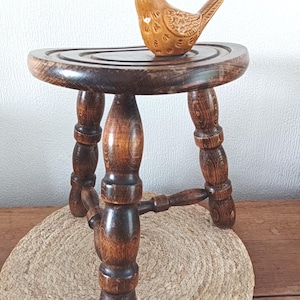 Peut inclure: Un petit tabouret en bois brun foncé doté de trois pieds tournés et d'une assise triangulaire arrondie. Posée sur l'assise, une petite figurine d'oiseau en céramique brun clair présente des détails d'ailes texturés et une finition lisse et brillante.