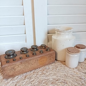 Peut inclure: Un bloc de bois rectangulaire contenant un ensemble de poids en laiton gradués de tailles variées, accompagné de trois pots en céramique blanc cassé, dont deux sont munis de petits bouchons en liège, disposés sur une surface tressée.