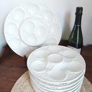 Könnte beinhalten: Weiße Austernplatten aus Keramik mit muschelförmigen Fächern. Ein Stapel Teller ist zu sehen, zusammen mit einem größeren Teller und einer Flasche Champagner. Die Teller sind zum Servieren von Austern und anderen Meeresfrüchten bestimmt.
