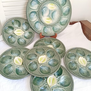 May include: A collection of seven round green ceramic oyster plates featuring molded shell-shaped indentations and a central raised lemon wedge motif. The plates have a textured, rope-like border design and a glossy finish in varying shades of green.