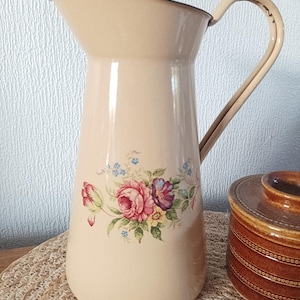 May include: Cream enamel jug with a floral pattern of pink, purple, and blue flowers. The jug has a curved handle and a dark brown rim. A brown ceramic jar is partially visible on the right.