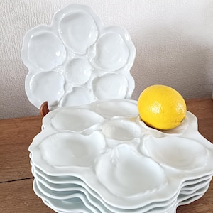Puede incluir: Un conjunto de platos de cerámica blanca para ostras, con múltiples compartimentos con forma de concha. Un limón amarillo entero descansa sobre uno de los platos. Los platos están sobre una superficie de madera.