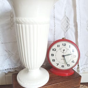 Puede incluir: Un jarrón alto de cerámica blanca con la parte superior acampanada y el cuerpo acanalado. Un despertador rojo con esfera blanca y números negros está junto al jarrón. El reloj tiene una base roja y una campana plateada.